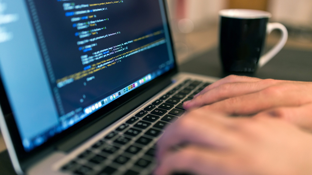 The hands of a coder programming on a laptop with a coffee cup in the background.